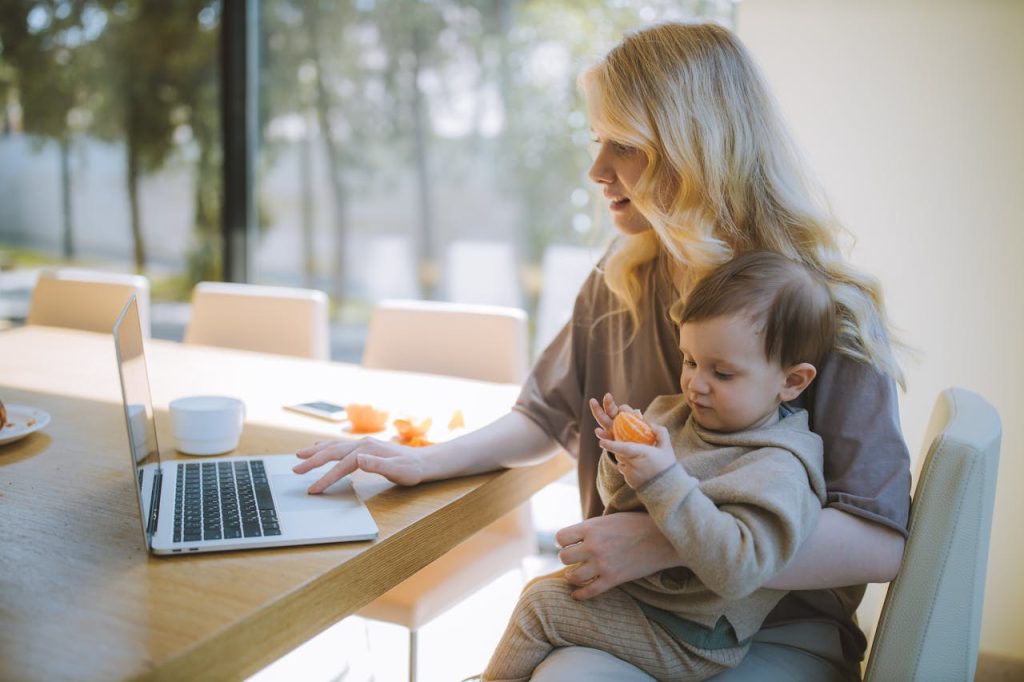 Una madre con capelli lunghi biondi è seduta al tavolo davanti al suo laptop tenendo in braccio il figlio